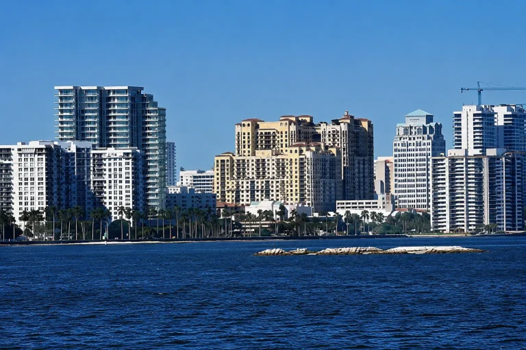 A city skyline with modern high-rise buildings is bordered by a body of water, with a small rocky island in the foreground.